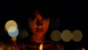 A woman looks intently at the camera with a blurred bonfire blazing behind her, flames dancing in the night.