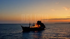 A fiery ship drifts across gilded waves at sunset, with the fire consuming the vessel and reflecting on the sea.