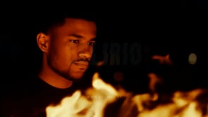 A man observes a bonfire with focused intent, the flames blurred in the foreground, creating a dramatic perspective.