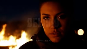 A close-up of a woman gazing intently as blurred bonfire flames fill the foreground, adding depth to the scene.