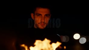 A man stands in contemplation, staring at a bonfire with flames prominently in the foreground, creating a dramatic visual.