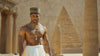 A muscular man from Ancient Egypt adorned in gold jewelry, with the majestic pyramids and ancient temples softly blurred in the background, emphasizing strength and opulence.